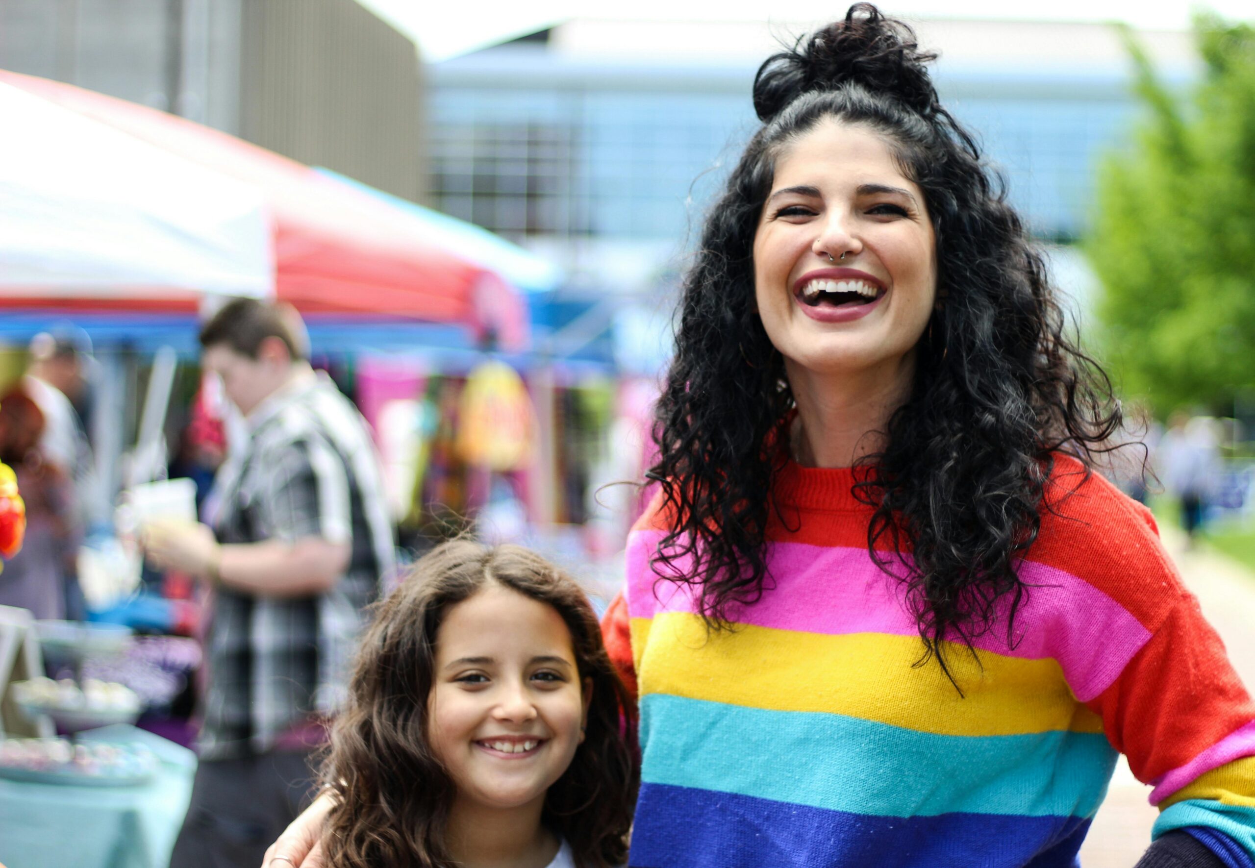 A mother and daughter share a joyful moment at a vibrant outdoor market fair.